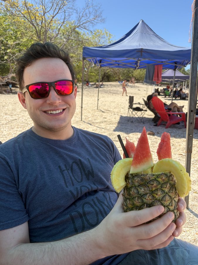Andrew smiling on a beach holding a pineapple drink